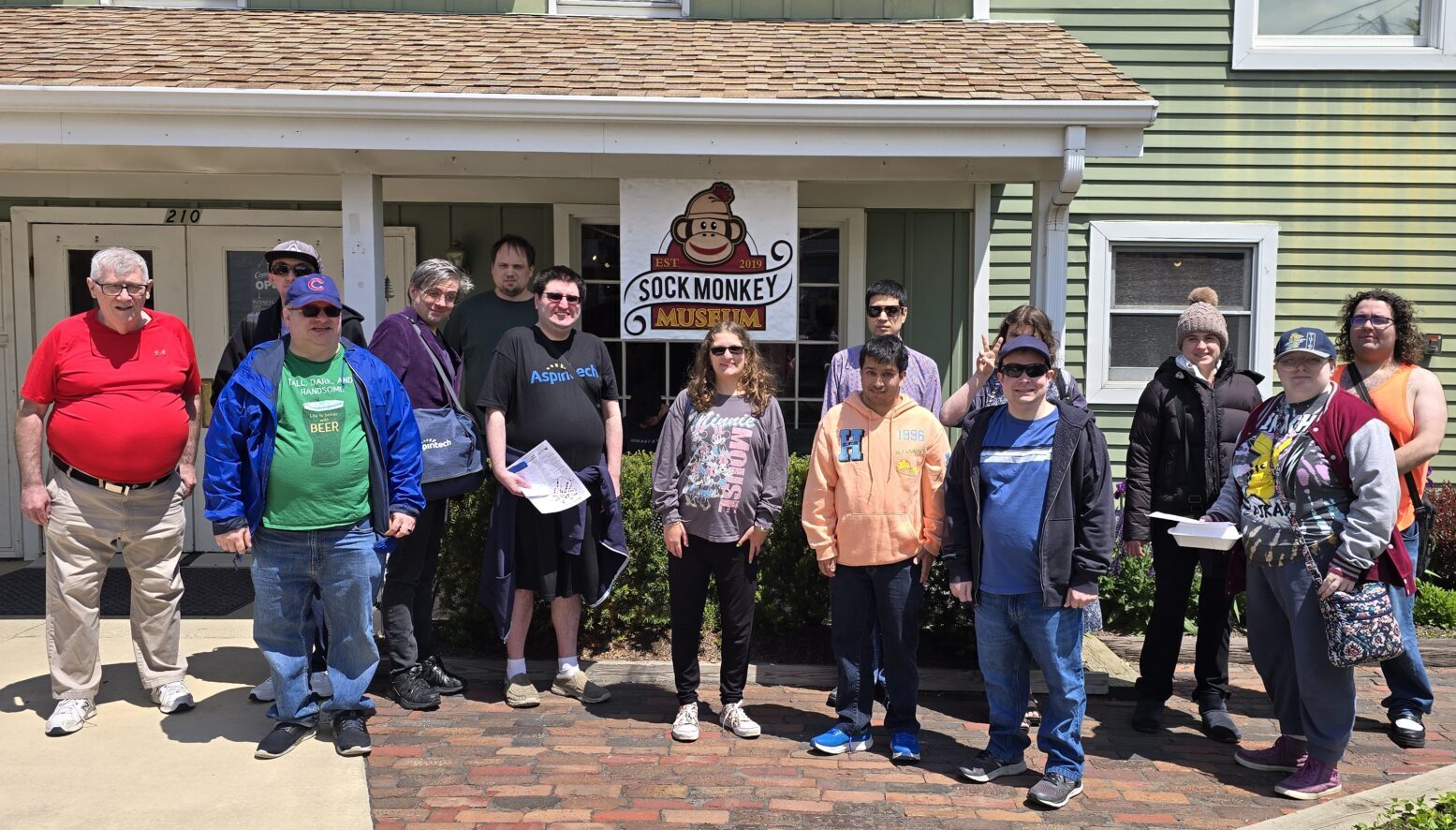 Fourteen Stepping Up & Out participants take a group photo in front of the sign for the Sock Monkey Museum, which was established in 2019. The museum is a light green painted building shown in the background.