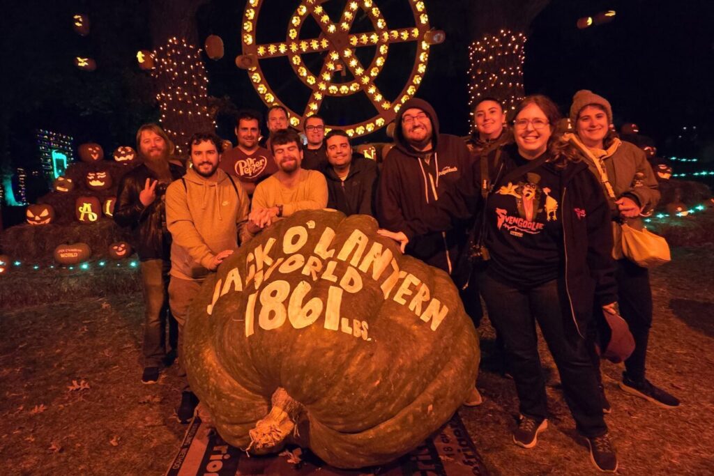 Eleven Stepping Up & Out participants take a group photo around an enormous pumpkin that has been carved to say "Jack O' Lantern World 1861 lbs." It is night time, with lights and additional pumpkin displays visible in the background.