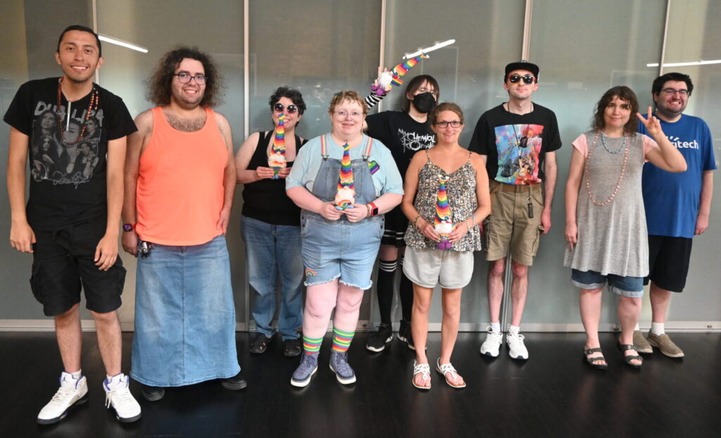 A group of nine SUO participants take a group photo in front of a glass wall at the Aspiritech office. Some are holding gnome dolls with rainbow hats.
