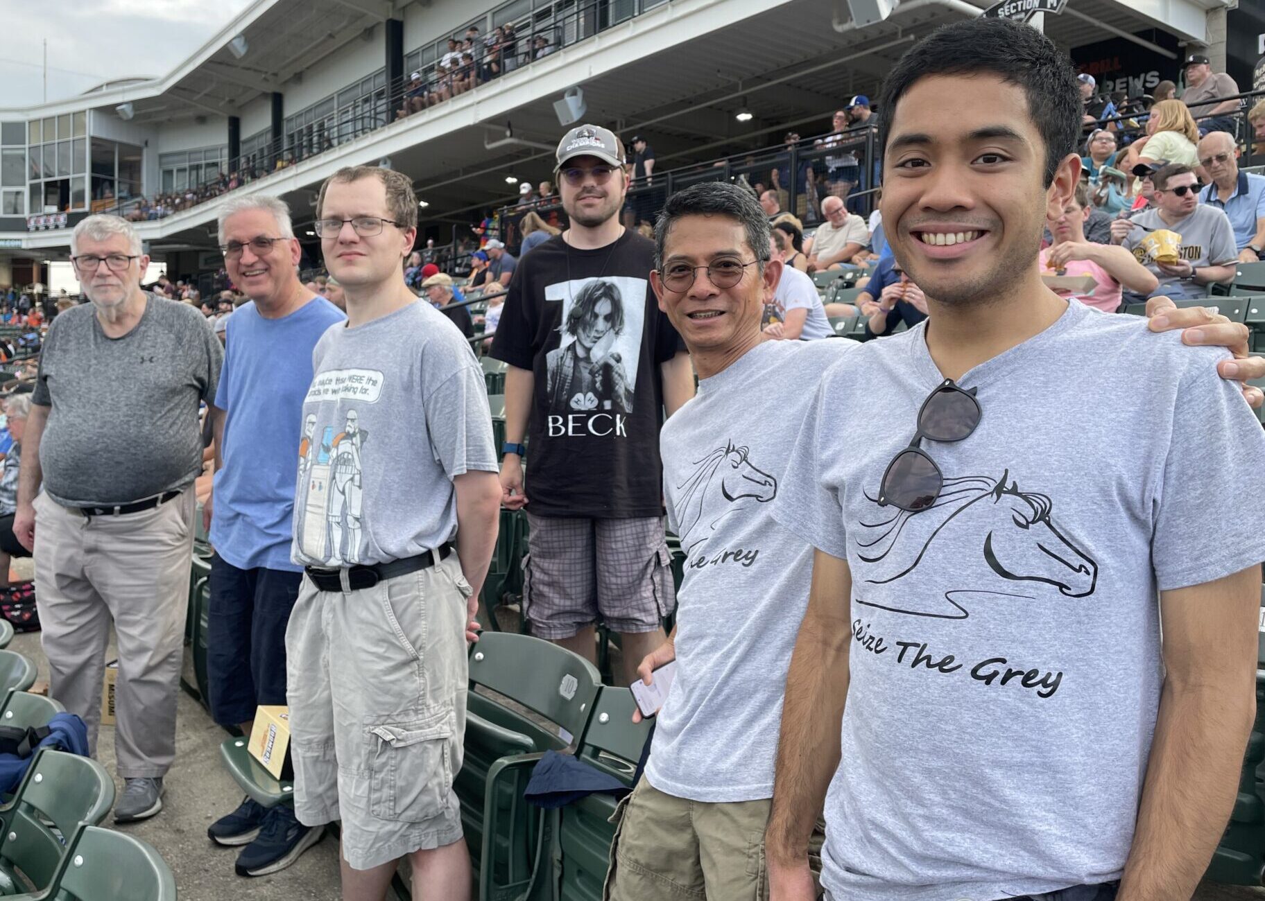 Six Stepping Up & Out participants stand for a photo in two rows of a stadium. A crowd of other event attendees is in the background.