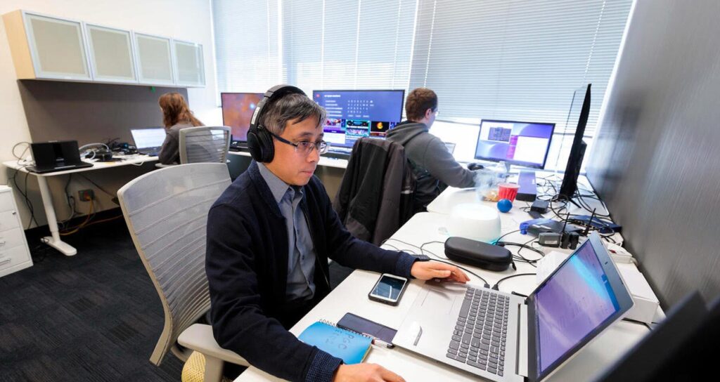 A male Aspiritech employee wears Bose headphones while testing them with a laptop and smartphone in an office.