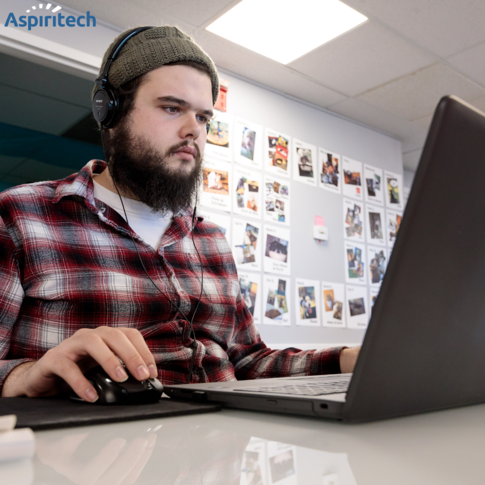 Aspiritech Data Services Program Manager Kyle Verbeke wears a red plaid shirt and green beanie while working at a laptop.