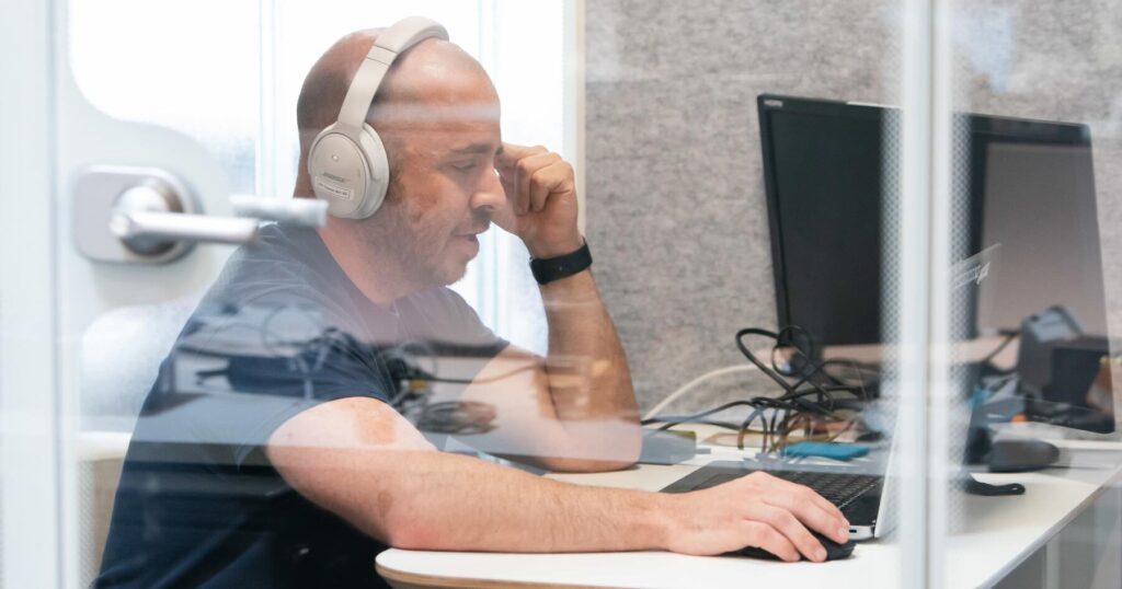 An Aspiritech employee wearing white headphones and a dark t-shirt works tests audio at a computer inside a clear reflective booth.