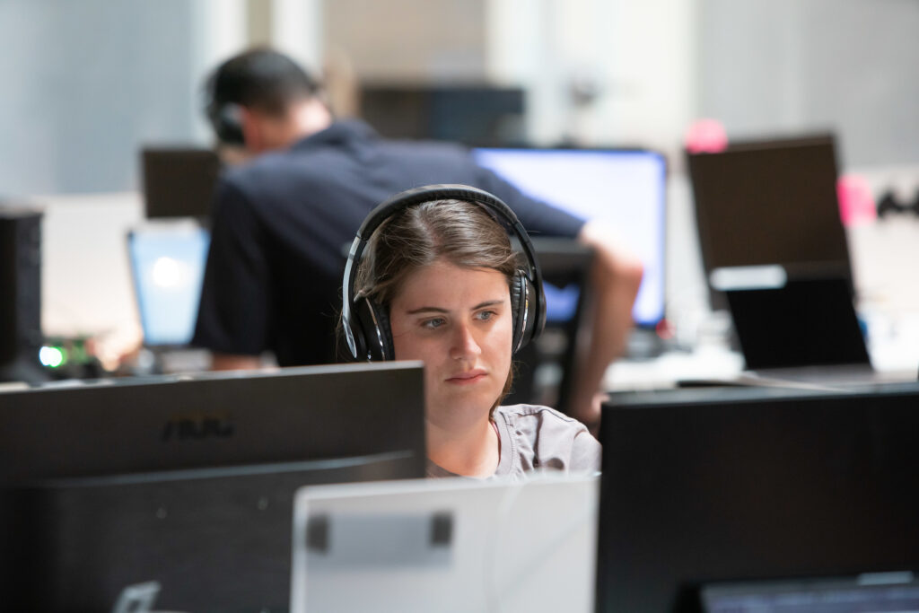 A female employee with brown hair wears headphones in front of a computer screen.