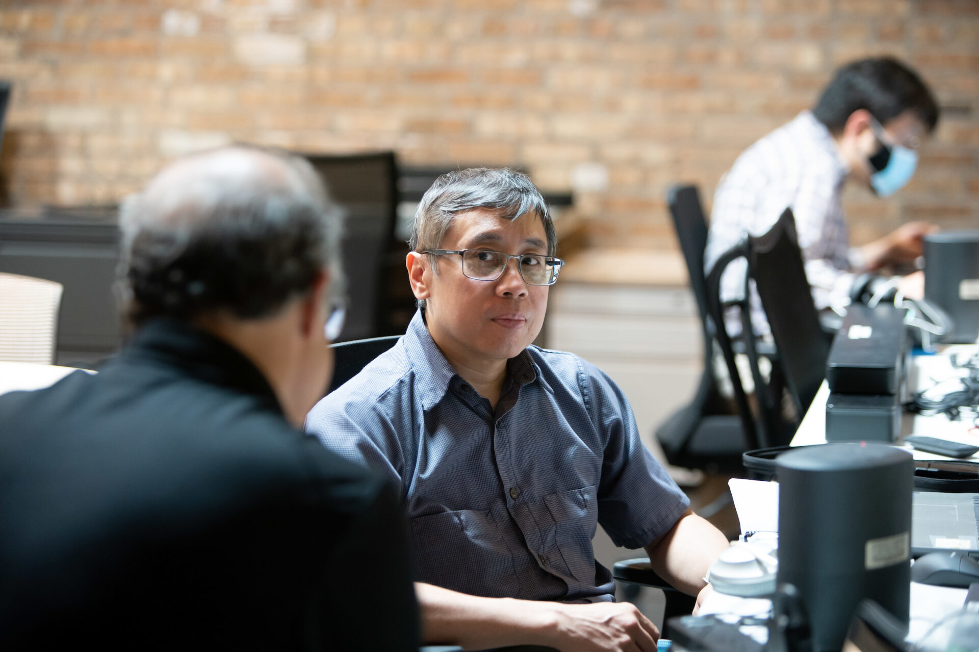 A male Aspiritech QA analyst in a blue-gray shirt and glasses looks toward a man in a black shirt in glasses.