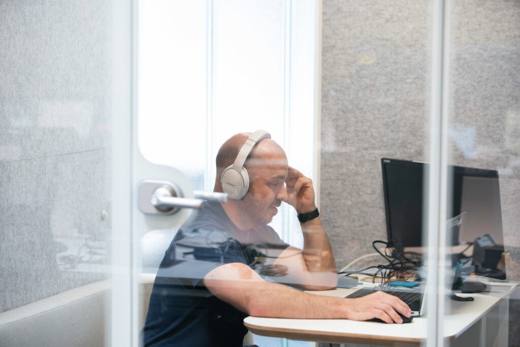 A male Aspiritech employee wearing white headphones tests audio at a computer inside a clear reflective booth.
