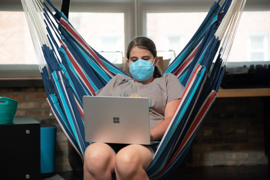 A woman wearing a mask works at a laptop in a hammock chair in an office.