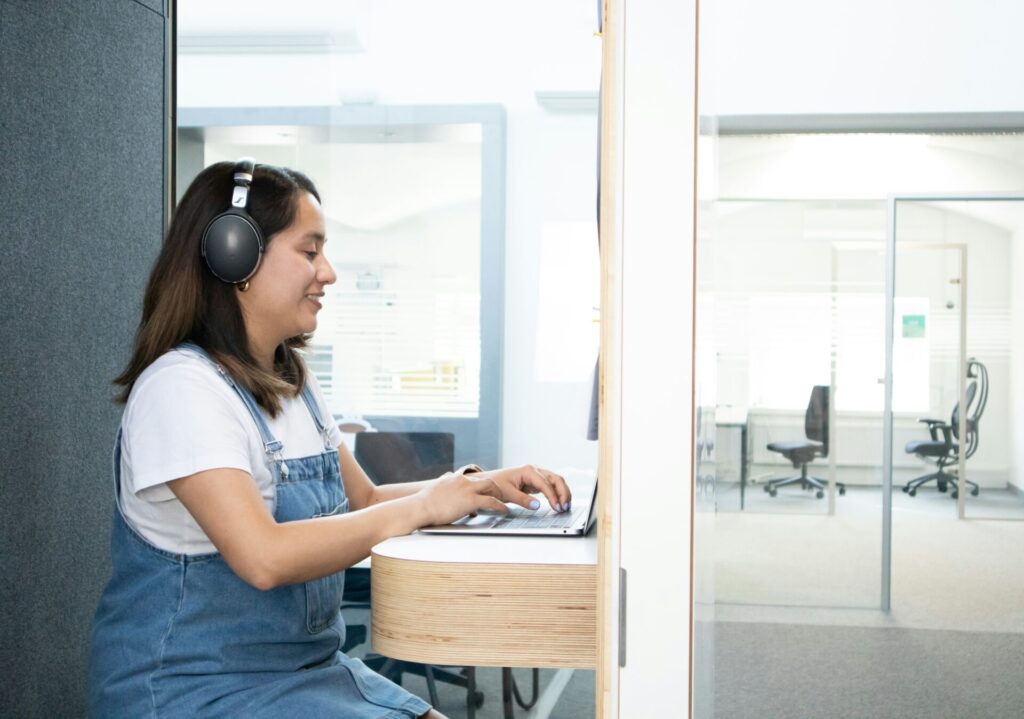 A dark-haired woman wearing headphones works at a laptop in a narrow booth.