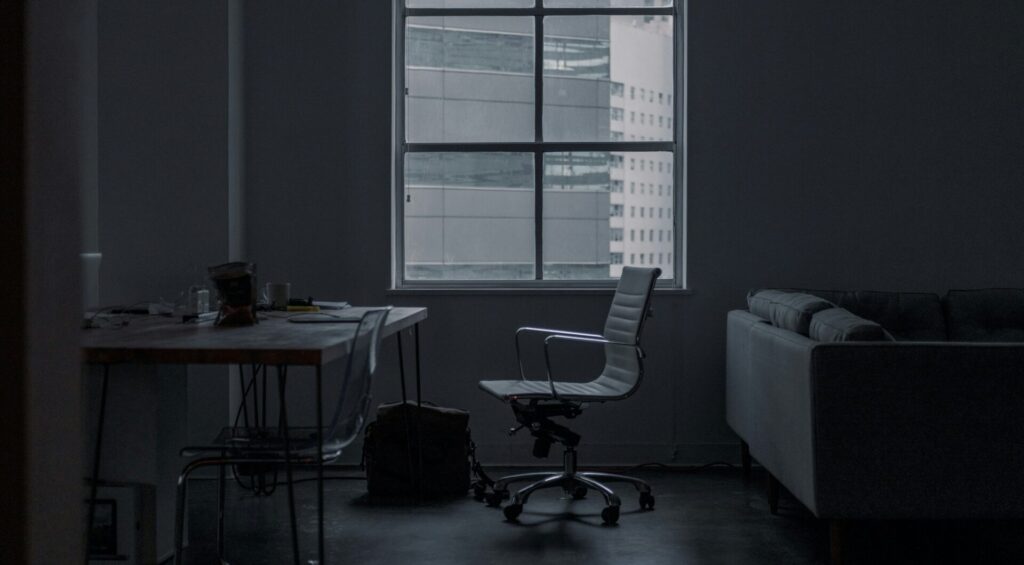 A desk chair sits empty in front of a window in a darkened room.