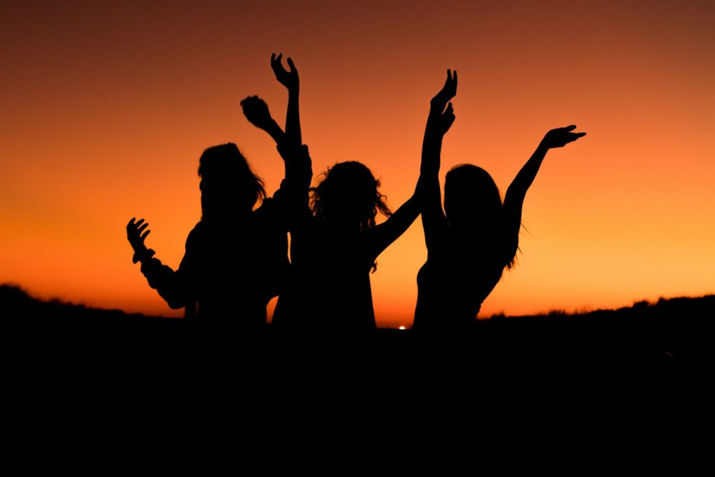 Three women stand in dark silhouette with arms upraised against an orange sunset background.