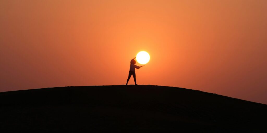 A silhouette of a person against an orange sky poses to look like they're holding the sun.