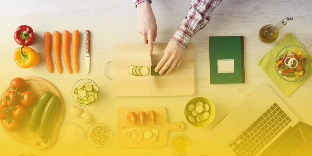 Hands hold a knife and slice a cucumber on a cutting board amidst an array of vegetables and a laptop.