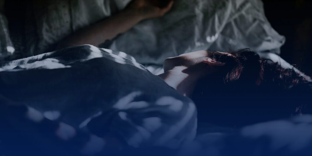 An adult person lies in a bed in a darkened room.