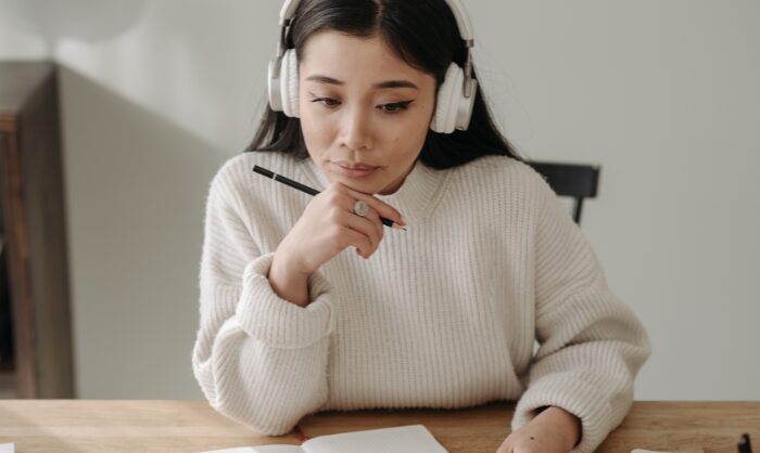 A young woman wearing headphones holds a pencil while studying an open book.