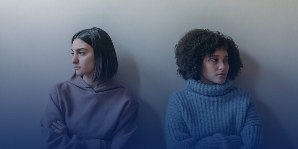 Two women stand against a blank wall, angrily looking away from each other.