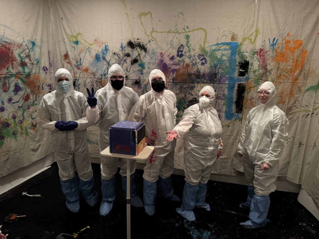 Five Aspiritech team members in white suits and masks stand in a room with white walls decorated with several splashes of color and handprints. One gestures at a small box sitting on a table.