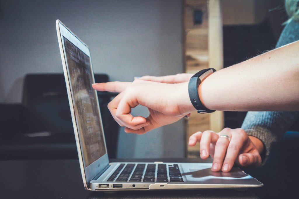 One person's hand points at a laptop screen while another person works the built-in mouse.