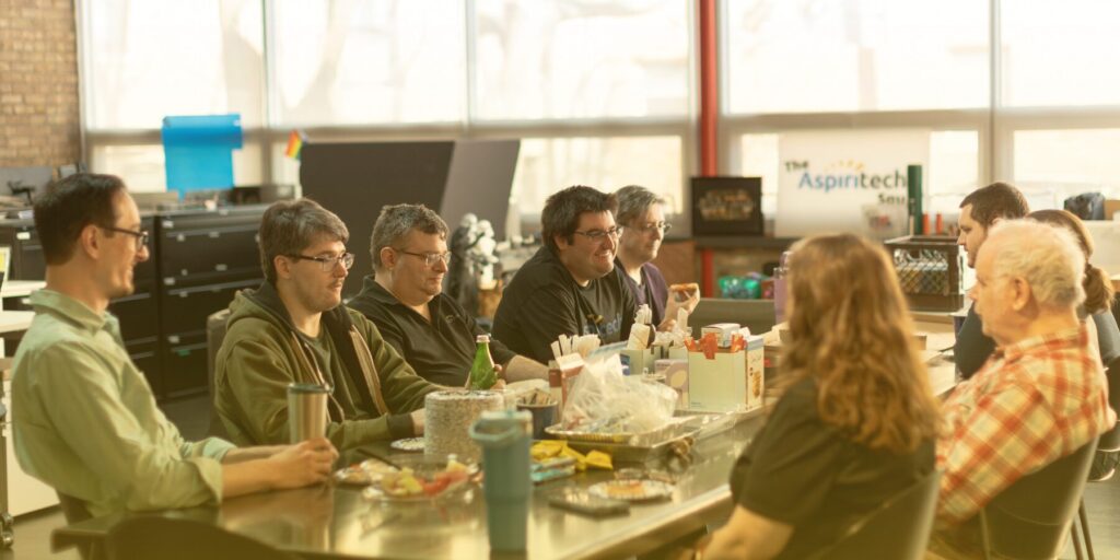 A group of nine Aspiritech employees talk over lunch on a long table.