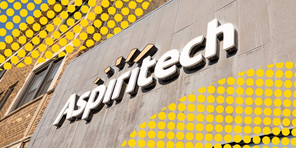 An angled look at the Aspiritech logo displayed on the exterior of the concrete exterior wall of the organization's headquarters. Yellow dot graphics overlay the top left and bottom right corners.