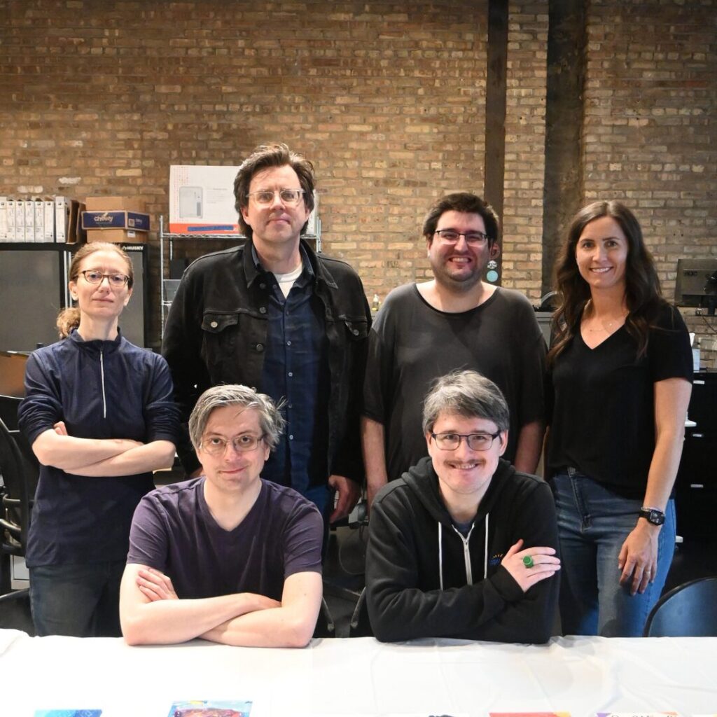 Six Aspiritech Creative Services team members and associates wearing dark clothes smile for a group photo in a brick-walled office, with four people standing in the back and two with folded arms sitting at a long table.
