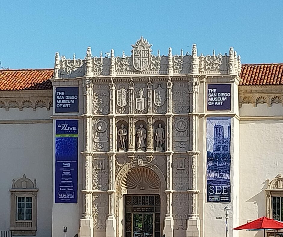SDMA Site Square An exterior view of the San Diego Museum of Art, with a detailed facade, blue posters, and a terra cotta tile roof against a clear blue sky.