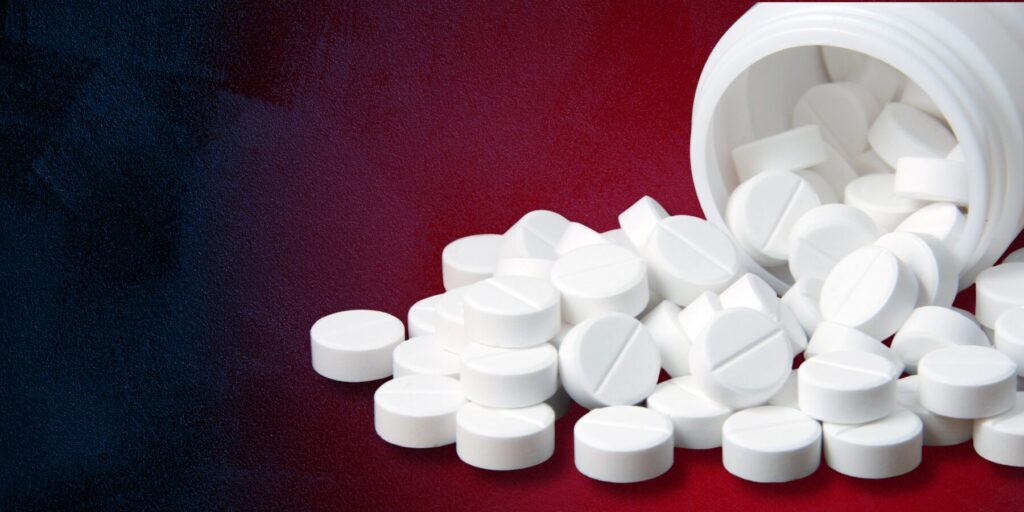 A spilled white bottle of white acetaminophen pills spotlit by a red glow against a deep navy textured graphic background.