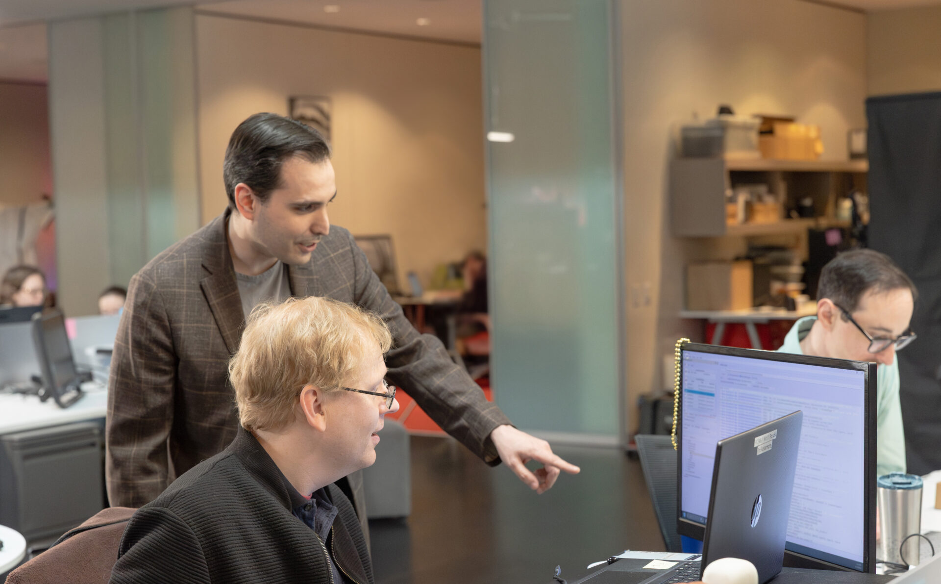 A male team leader in a brown suit jacket points at something on a screen while another male employee in a black zip-up sweater looks on. A third male employee in a light green shirt works at his station in the background.