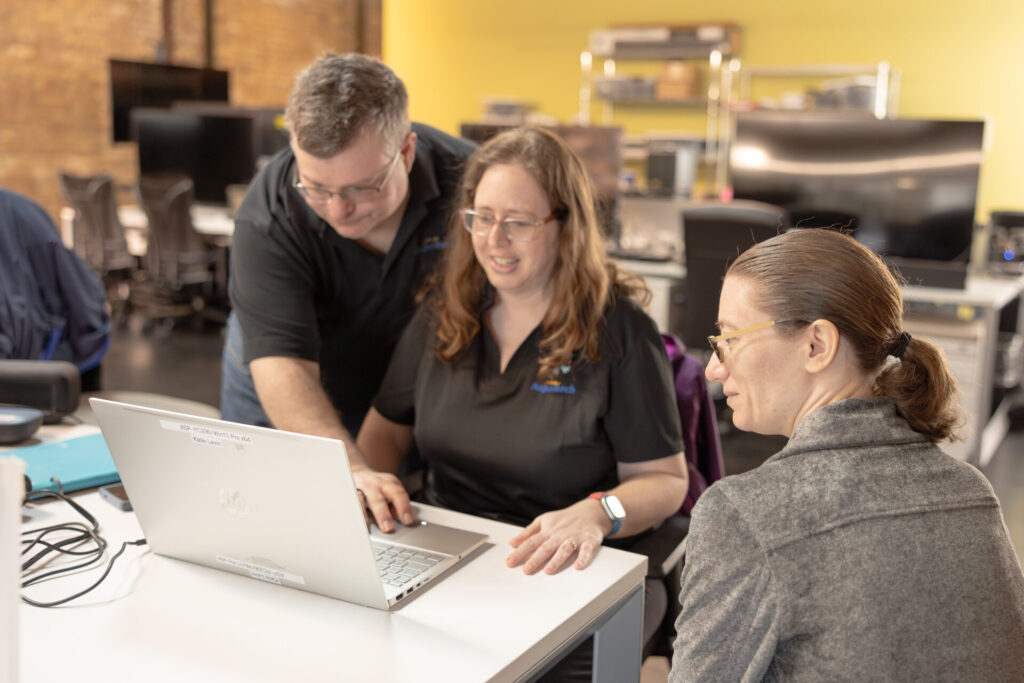 Two female employees sit by a laptop while a male employee reaches over to move the mouse to show something on the screen. They are in an open office with yellow and exposed brick walls.