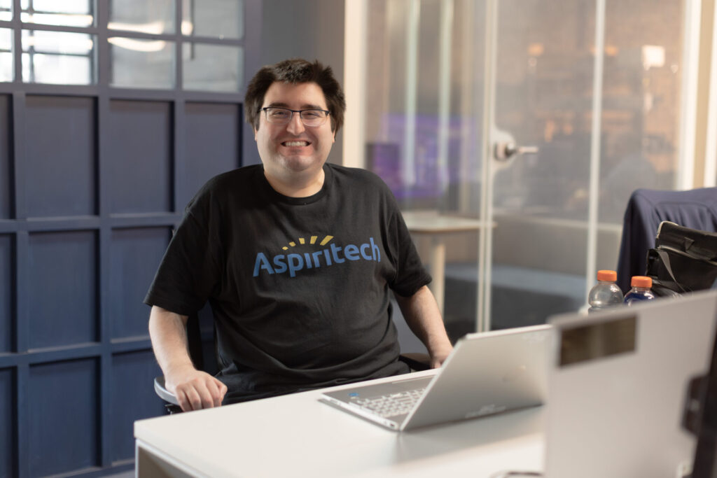 A male Aspiritech employee wearing a black Aspiritech t-shirt smiles at a laptop on a long white desk. He's in an open office setting with blue and clear glass booths in the background.