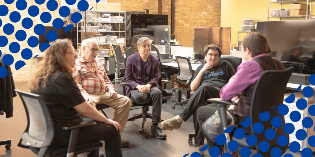 A group of five Aspiritech employees sit in a circle of office chairs, appearing to have an active discussion. They are in an open office setting with yellow and exposed brick walls. Graphic blue dots decorate two corners of the image.