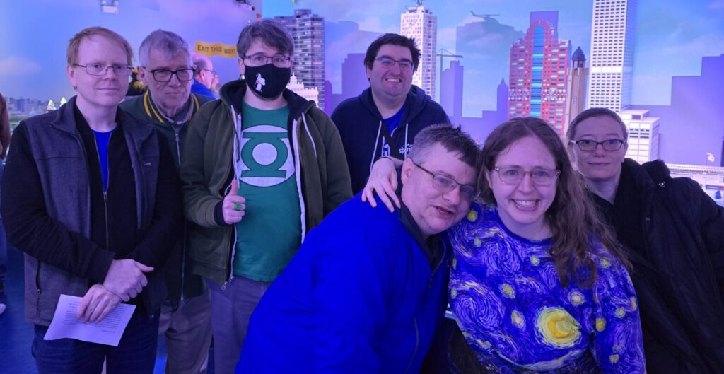 A group of seven Stepping Up & Our participants take a group photo against a skyline made out of LEGO bricks.