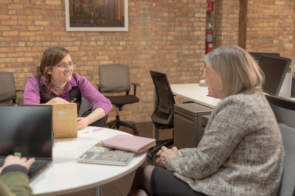An Aspiritech employee wearing a purple shirt and vest talks at a table with the CEO in an open office setting with exposed brick walls.