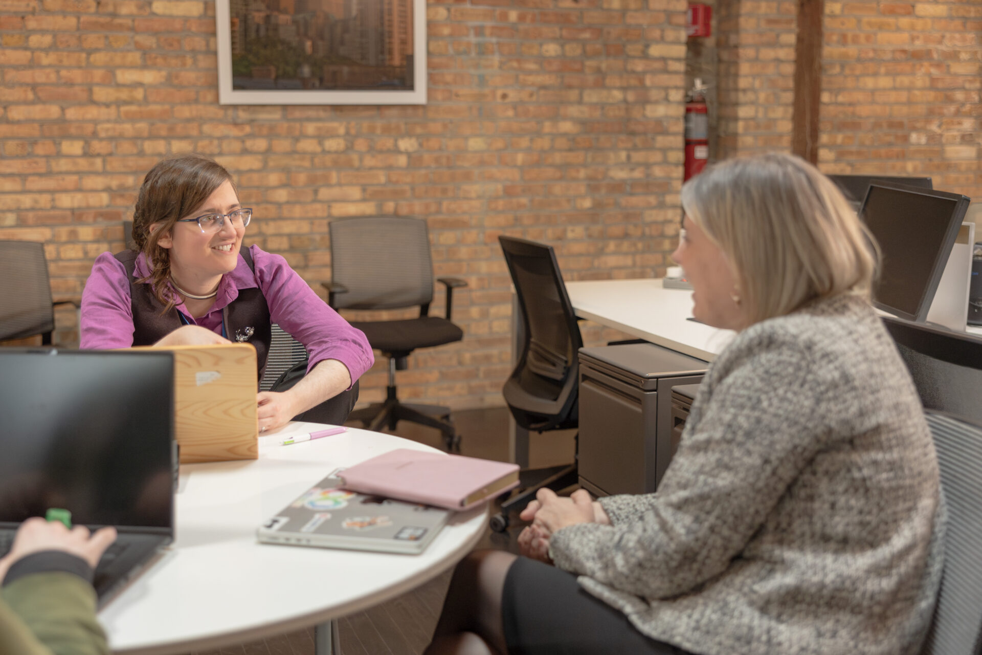 An Aspiritech employee wearing a purple shirt and vest talks at a table with the CEO in an open office setting with exposed brick walls.