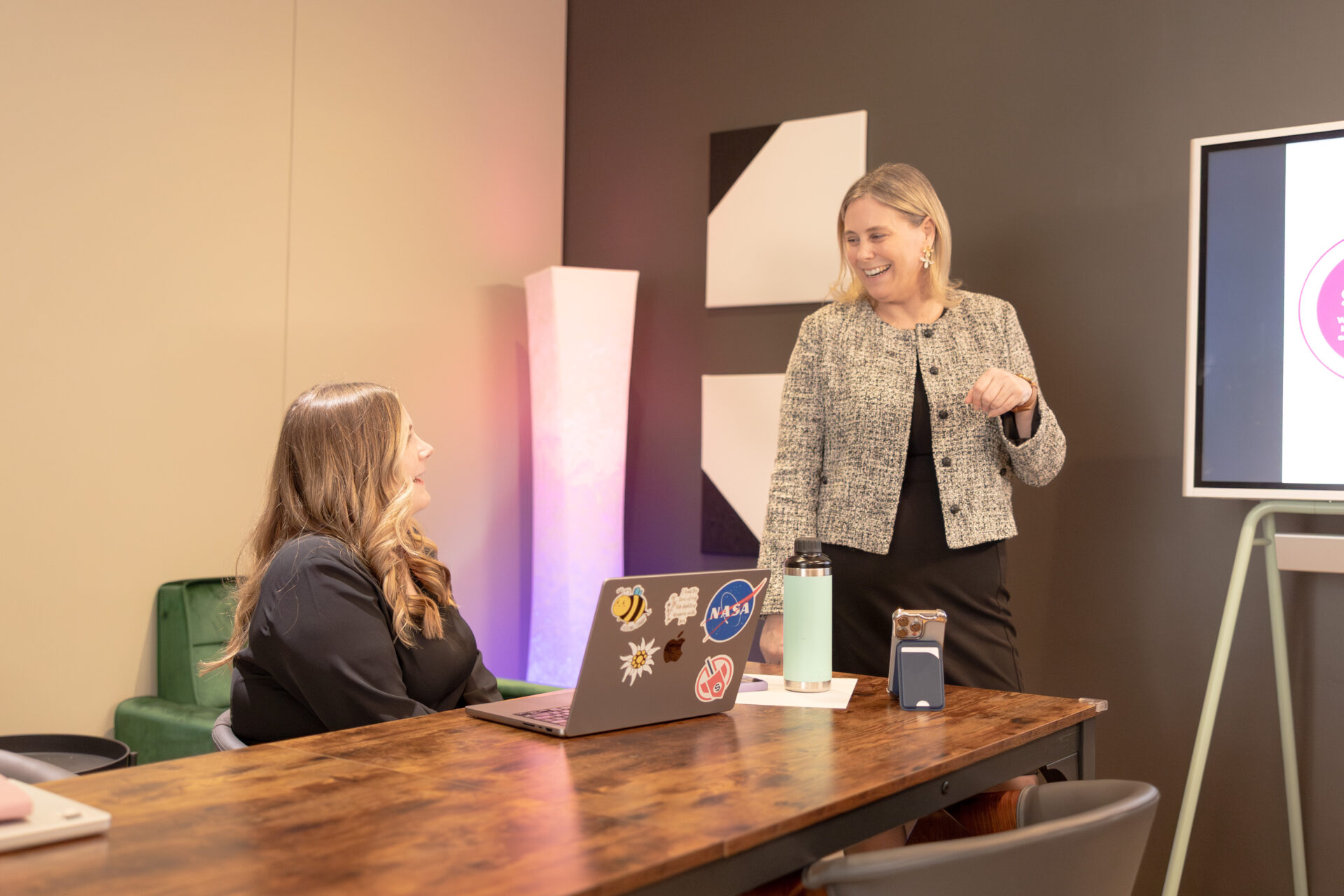Two female Aspiritech executives talk in a conference room decorated with a green velvet sofa, purple ambient light, and modern geometric paintings. One sits at a wood table with her laptop while the other stands.