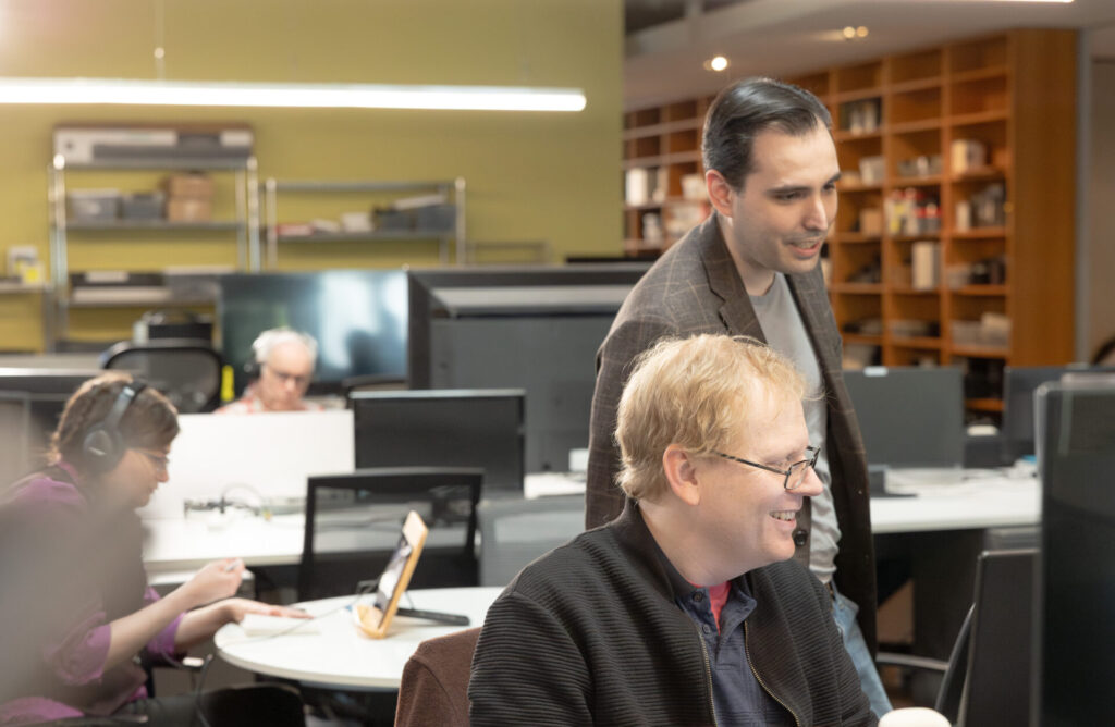 A male Aspiritech team member stands and looks over a second seated team member's shoulder. They're viewing something on a computer screen.