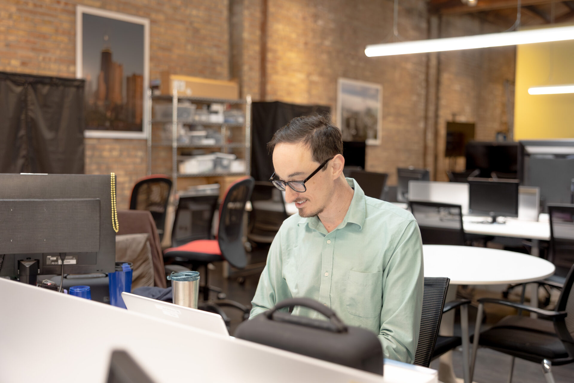 A male Aspiritech employee wearing glasses and a light green shirt works at a laptop in an open office setting.