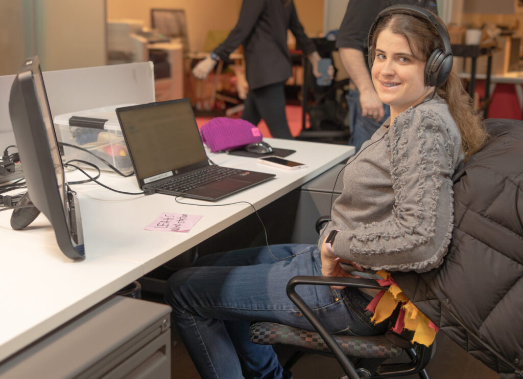 A female Aspiritech employee wearing a gray sweater and headphones smiles at the camera. She's sitting at her workspace in an open office, with additional employees passing by in the background.