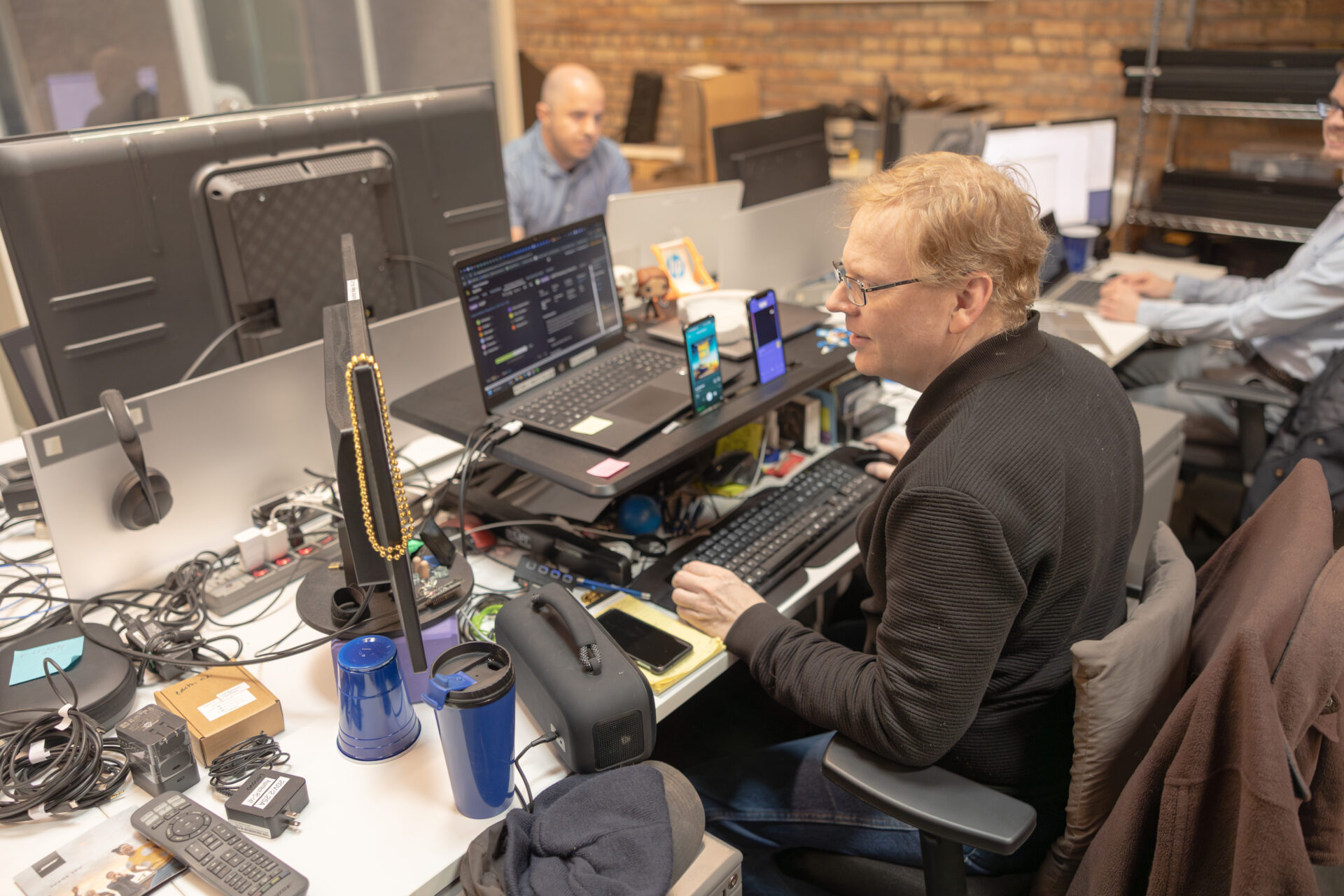 A male Aspiritech employee wearing a black sweater and glasses sits at a workstation featuring a raised laptop, computer monitor, three smartphones, and other items.