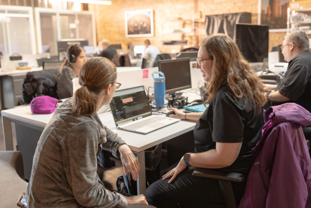 Two female Aspiritech employees look at a laptop screen while sitting at the corner of a long white desk in an open office setting.