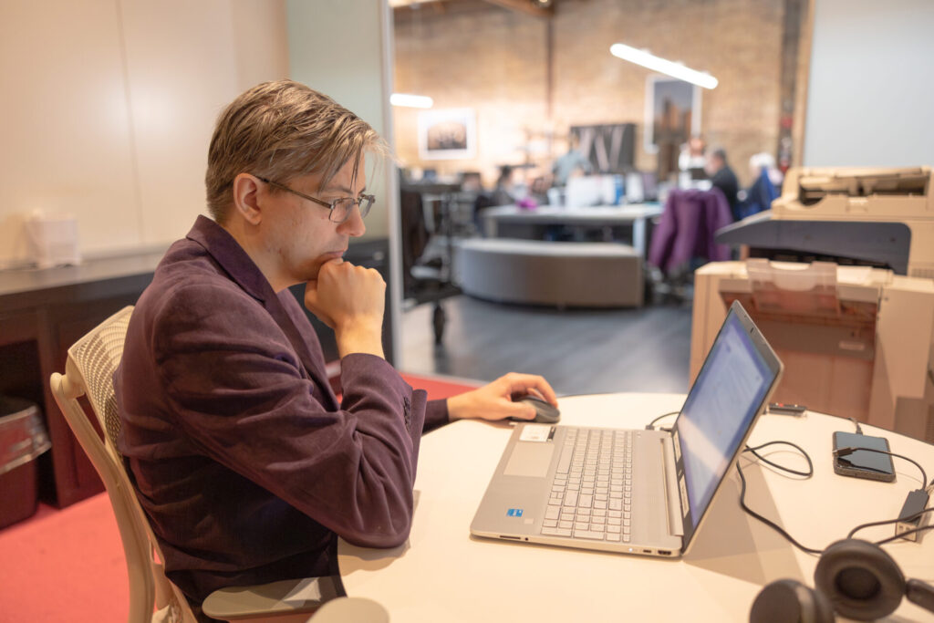 A male Aspiritech employee wearing a burgundy jacket and glasses works at a laptop.