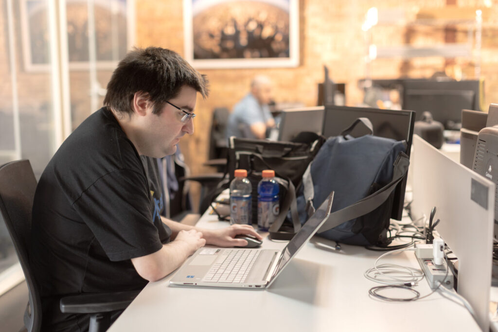 A male Aspiritech employee wearing a black shirt and glasses works at his laptop on a long white desk in an open office with exposed brick walls.