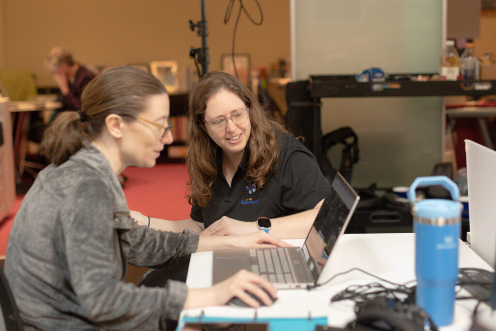Two female Aspiritech employees smile while looking at a laptop screen.