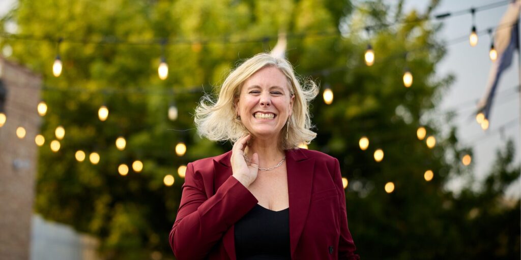 Tara May gives a big smile while wearing a red blazer and black blouse. Green foliage and strings of light are shown in the background.