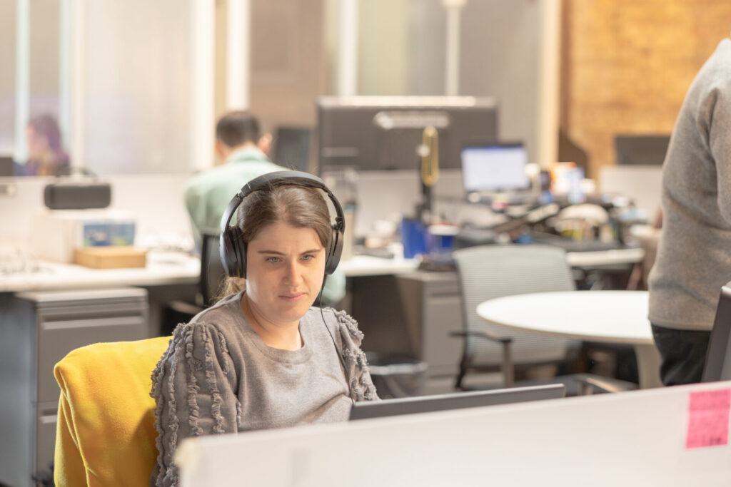 A female Aspiritech employee wearing headphones and a gray sweater looks at a computer screen in an open office setting.