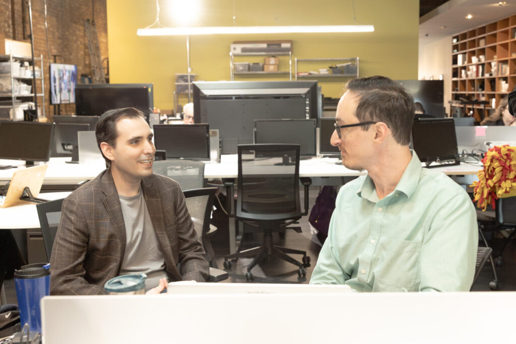Two male Aspiritech employees sit talking at a desk in an open office.