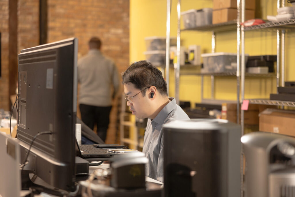 An Aspiritech employee sits at a workstation with a laptop, a large monitor, and two smartphones.