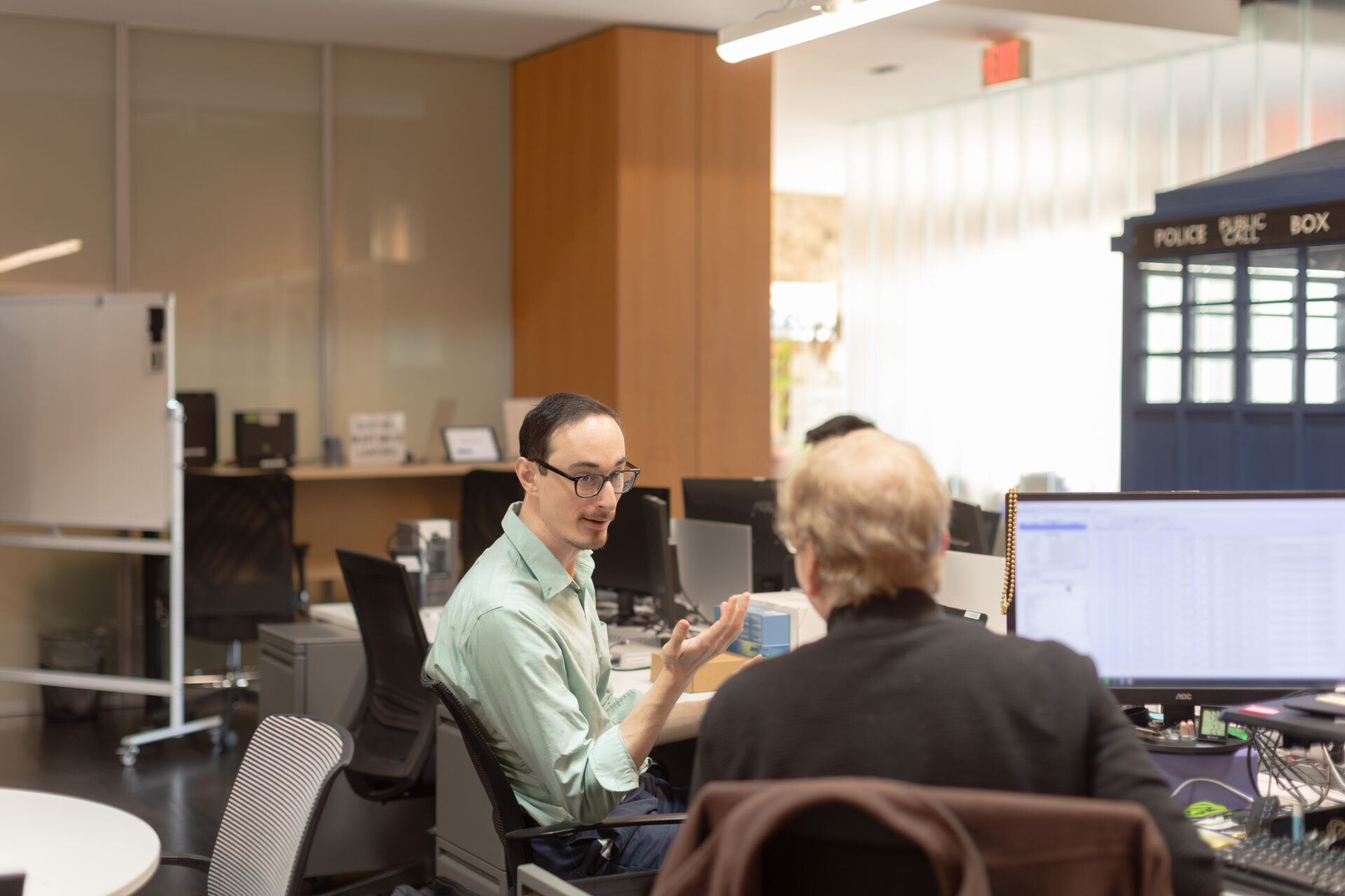 A male Aspiritech employee explains something to another employee as they both sit at computer workstations.