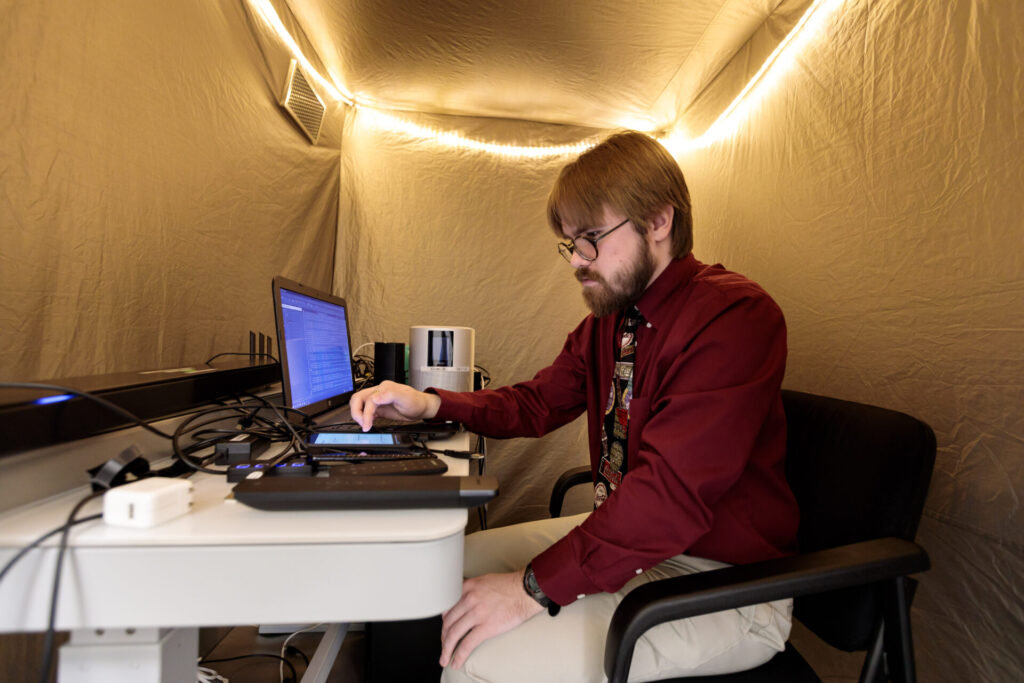 A male Aspiritech employee tests speakers in a sound booth equipped with string lights.