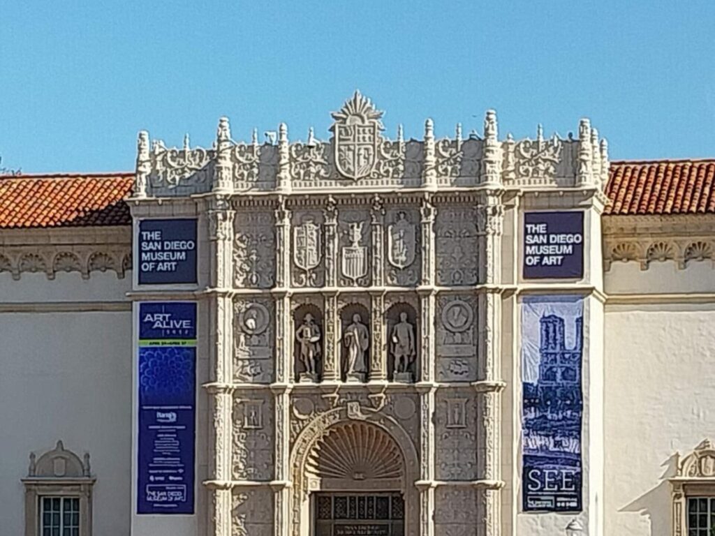 An exterior view of the San Diego Museum of Art, with a detailed facade, blue posters, and a terra cotta tile roof against a clear blue sky.