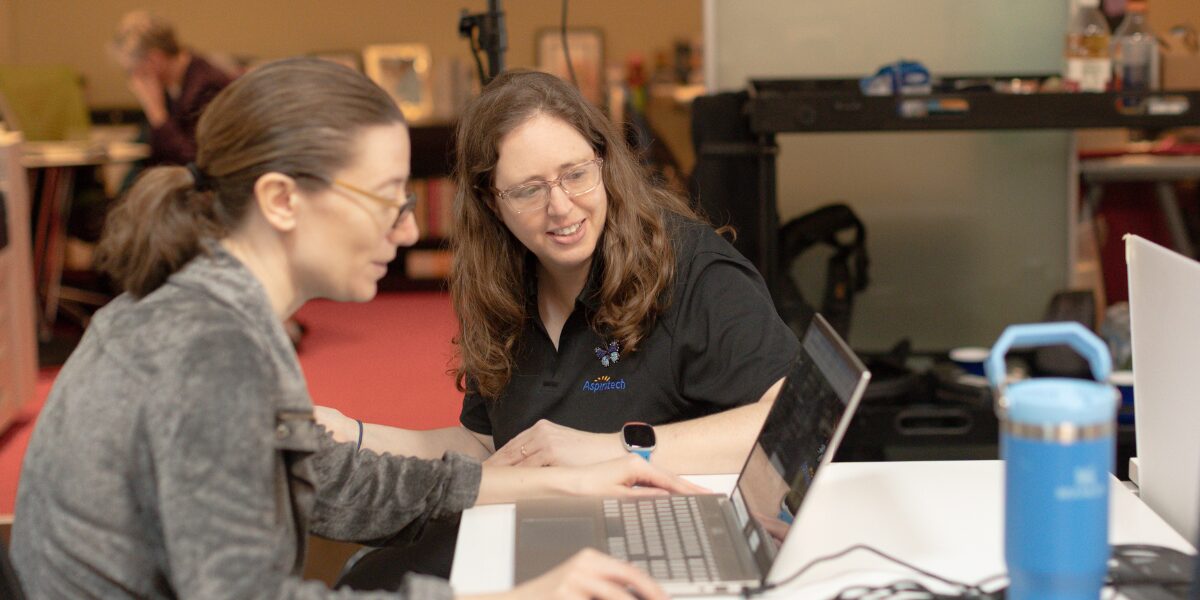 Two female Aspiritech employees smile while looking at a laptop at a workstation.